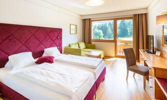 A modern hotel room with a large double bed and a green couch. The room has large windows with a view of a green landscape.