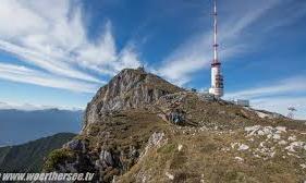 A rocky mountain with a transmission tower and far-reaching views. The sky is blue with many clouds.