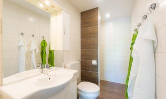 A modern bathroom with white tiles and a large mirror. Clean towels hang on the wall, and there is a bright shower area.