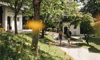 A couple is strolling leisurely through a green garden landscape with trees and flowers. In the background, some charming buildings can be seen.