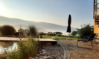 A tranquil garden with a walkway and a small body of water. In the background, gentle hills and a clear sky can be seen.