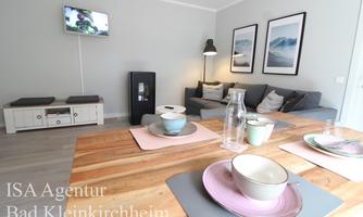 A modern kitchen with a dining table and colorful dishes. In the background, there is a cozy living room with a TV and a picture on the wall.