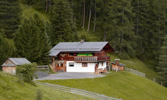 A rustic cabin surrounded by green meadows and trees. In the background, gentle hills are visible.