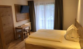 A cozy bedroom with a double bed, wooden details, and a table with chairs. The room has large windows that let in plenty of light.