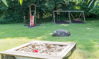 A playground with a sandbox and a slide can be seen. In the background, there are a swing and a lot of green grass.