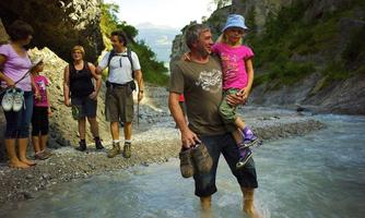 A group of people is hiking through a flowing stream in a picturesque gorge. A father is carrying his child, while others are enjoying nature.