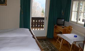 A simple room with two beds and a desk. The balcony offers a view of nature and the windows are adorned with green curtains.