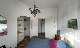 A bright bedroom with a large window and a beautiful chandelier. The walls are painted in neutral colors, and there is a shelf as well as a wardrobe.