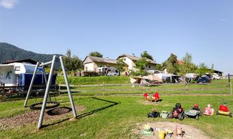 A playground with swings and colorful toys on a green meadow. In the background, motorhomes and houses can be seen, surrounded by trees.