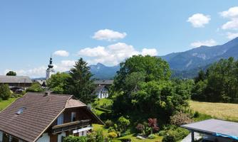 A picturesque mountain landscape with a village and a church in the foreground. Well-kept gardens and green meadows can be seen in the front.