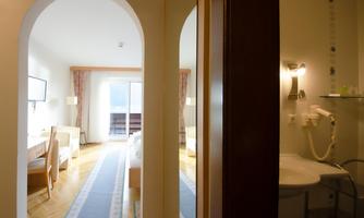 A well-designed hotel room with a wooden table, a television, and access to a balcony. In the background, a modern bathroom is visible.
