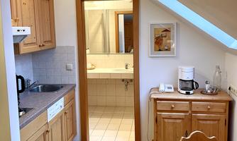 A cozy kitchen area with wooden cabinets and a dining area. In the background, a bathroom is visible through a door.