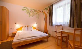 A cozy room with a double bed and a wooden table. A beautiful tree is painted on the wall, and large windows provide plenty of daylight.