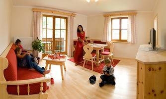 A cozy living space with a light wooden floor and large windows. A family is sitting together while a woman in a red dress stands.