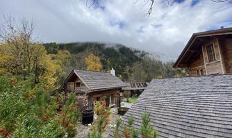 A tranquil landscape with traditional wooden houses and colorful autumn foliage. In the background, wooded hills and a cloudy sky can be seen.