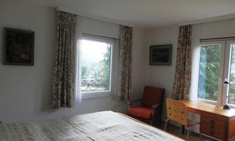 A cozy bedroom with a large bed, a desk, and a chair. The windows offer a view of the outdoors, surrounded by curtains with floral patterns.