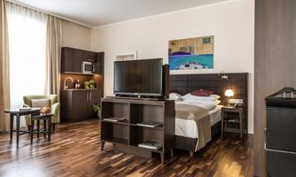 A modern hotel room with a double bed, a television, and a small kitchenette. The room is bright and friendly with wooden flooring and a piece of art on the wall.