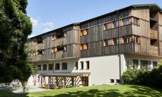 A modern building with a wooden facade and large windows. It is situated in a green environment with lawns and trees.