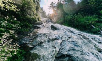 A glittering waterfall flows over smooth rocks. Surrounded by lush greenery and trees, the sun shines through the clouds.
