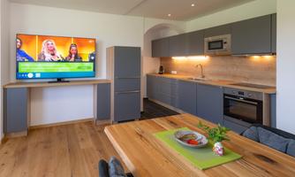 A modern kitchen with gray cabinets and a wooden countertop. Below is a dining table with a decorative plate and a television on the wall.