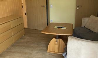 A bright room with a round wooden table, a comfortable armchair, and a dresser. Two doors and a mirror hang on the wall.