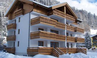 A modern building in alpine style with wooden balustrades and several balconies. It stands in a snowy landscape under a clear sky.