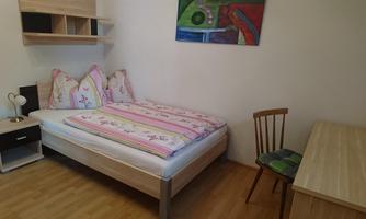 A simple bedroom with a double bed and colorful cushions. A table and a chair complement the space, which appears bright and friendly.