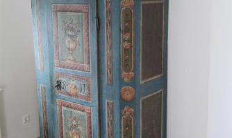 An antique, blue-painted cabinet with colorful decorations stands in a bright room. On top of the cabinet is a pot with a flowering plant.