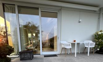 A modern outdoor area with a table and two chairs. Large windows and plants are visible in the background.