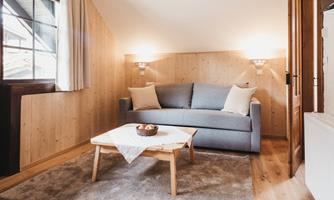A cozy living room with a gray sofa and a wooden table. The walls are made of wood and there is plenty of natural light.