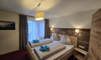 A cozy bedroom with two beds and modern wood details. The room has large windows and is brightly lit.