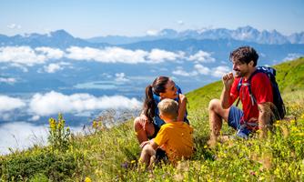 A family is sitting on a green meadow, enjoying the view of the mountains. They laugh and chat as clouds float over the valley.
