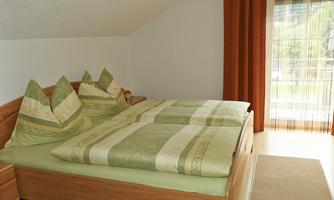 A cozy bedroom with a double bed and green pillows. Large windows let in plenty of daylight and offer a view outside.