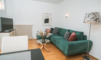 A modern living room with a green sofa and colorful patterned cushions. The room is bright and spacious, with a table and a television.