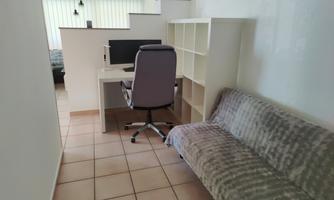 A modern room with a desk and a chair. In the corner is a gray sofa, and the light comes in through large windows.