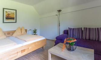 A cozy room with a double bed and a purple sofa. Next to a table is a plant in a glass vase.
