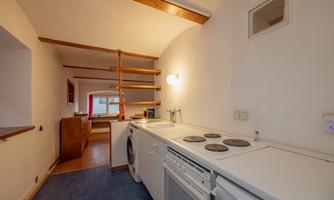A small kitchen with a bright work surface and modern appliances. There is an open shelf and windows that provide natural light.