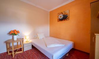 A simple room with a single bed and a bedside table. A painting hangs on the wall, and a small table with flowers stands next to it.