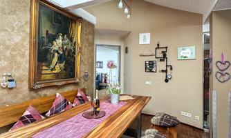 An inviting dining area with a large wooden table and comfortable seat cushions. A piece of artwork hangs on the wall, and the windows provide plenty of light.