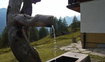 A wooden sculpture with a water feature stands in a green landscape. In the background, mountains and trees can be seen.