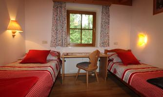 A cozy bedroom with two single beds and red cushions. There is a table with a chair and a window that lets in plenty of daylight.