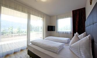 A modern bedroom with two beds and large windows. The room is bright and inviting, overlooking the nature outside.