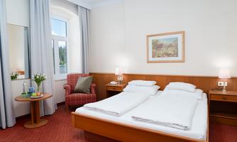 A bright hotel room with a double bed and a cozy armchair. There is a table with flowers and decorative objects.