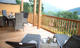 A cozy terrace with a wooden floor and a view of the green landscape. There are chairs, a table, and some drinks on the terrace.