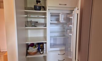 A refrigerator with an open door shows some empty shelves and dishes. Next to the refrigerator stands a shelf with various kitchen utensils.