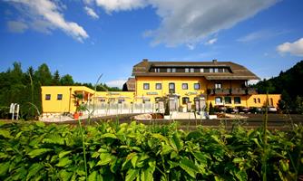 A charming hotel with a yellow facade, surrounded by green plants. The sky is blue with some white clouds.