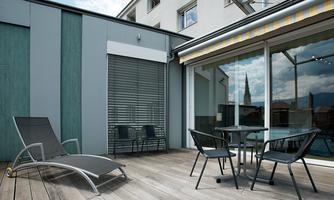 A modern terrace with a wooden floor and a table with chairs. In the background, windows and a view of the surroundings can be seen.