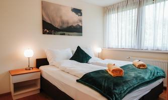 A cozy bedroom with a large double bed. The walls are light, and there are large windows that let in plenty of light.