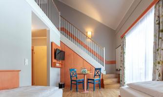 A bright and modern room with two beds and a staircase. A table with two blue chairs stands in front of a large window.