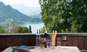 A table with drinks and cutlery, surrounded by a picturesque landscape with mountains and a lake. The view is green and inviting, ideal for a relaxing meal.